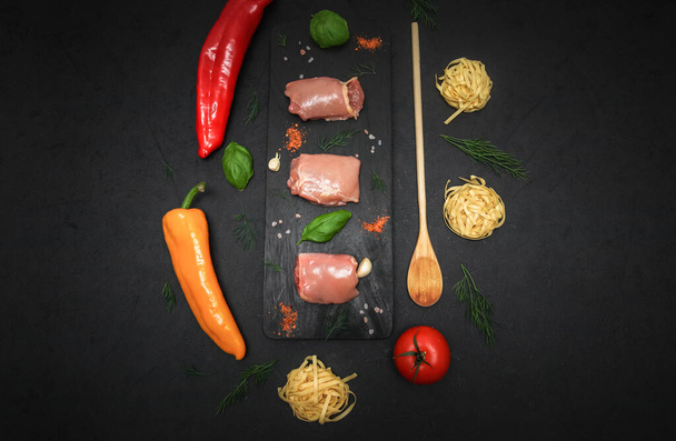 Raw chicken thighs on a black wooden board with spices, herbs, yellow peppers and fresh noodles lie in the middle on a black background, flat lay close-up. Cooking food concept. - Photo, Image