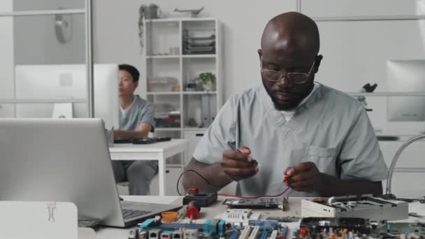 African American engineer testing electronic component with multimeter and working on laptop at desk in laboratory - Footage, Video