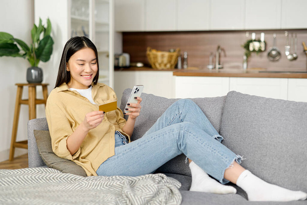 Asian woman consumer holding credit card and smartphone sitting on sofa at home making online banking transaction - Foto, immagini
