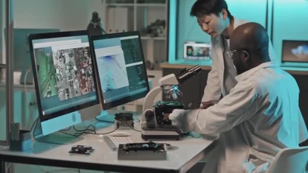 Tilt up shot de ingenieros técnicos multiétnicos en batas blancas discutiendo imágenes en la pantalla del ordenador conectada al microscopio mientras se examina el componente electrónico en el laboratorio - Metraje, vídeo