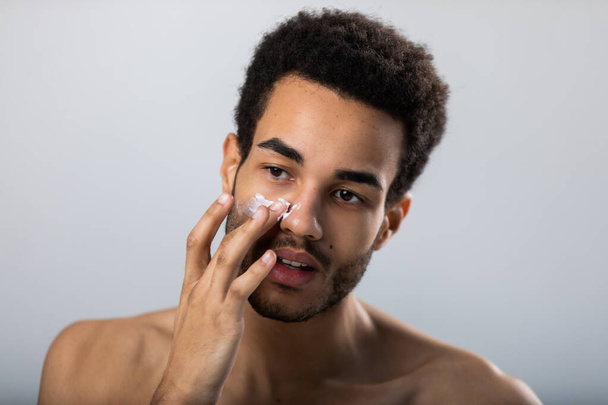 Young african american man applies moisturizer on his face. Male skin care. - Фото, зображення
