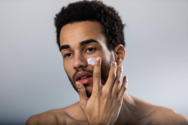 Young african american man applies moisturizer on his face. Male skin care. - Foto, Imagem