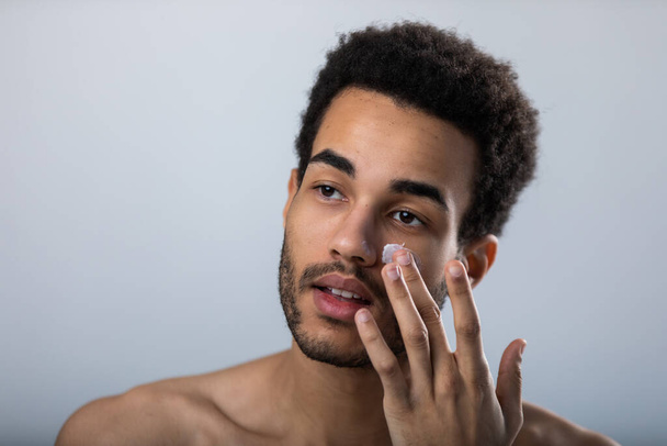 El joven afroamericano se aplica crema hidratante en la cara. Cuidado de la piel masculina. - Foto, Imagen