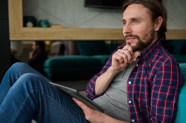 Pensive handsome bearded Caucasian man in casual denim and checkered shirt holding a digital tablet and looking thoughtfully away, sitting at home environment - Photo, image
