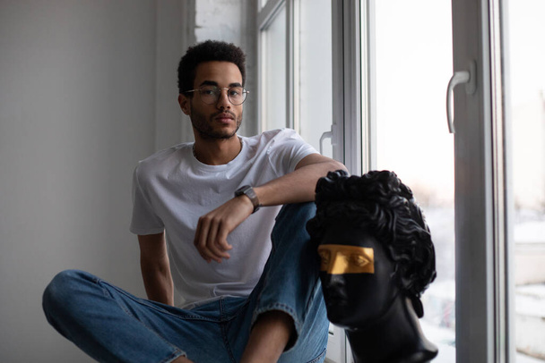 Attractive african american man in glasses and a white t-shirt sits on a windowsill. - Foto, Imagen