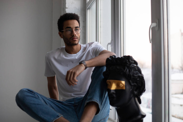 Attractive african american man in glasses and a white t-shirt sits on a windowsill. - Φωτογραφία, εικόνα
