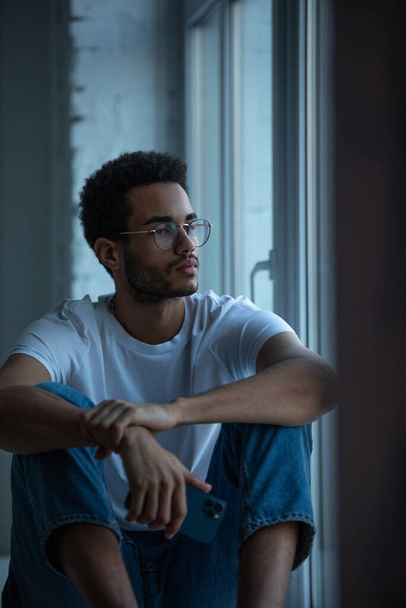 Attractive african american man in glasses and a white t-shirt sits on a windowsill. - Photo, image