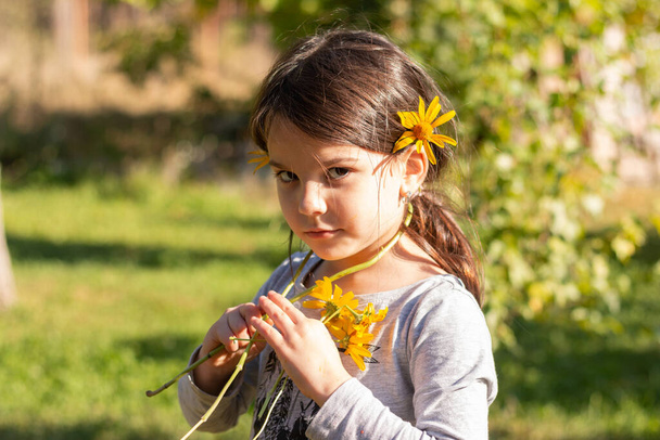 Une petite belle petite fille préscolaire triste avec des fleurs jaunes dans ses cheveux se tient à l'extérieur sur un fond d'été flou Une petite belle fille préscolaire triste avec des fleurs jaunes dans ses cheveux se tient à l'extérieur sur un fond d'été flou - Photo, image