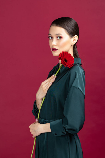 Lovely girl wearing green dress standing isolated over red background, posing with a herbera flower - Photo, Image