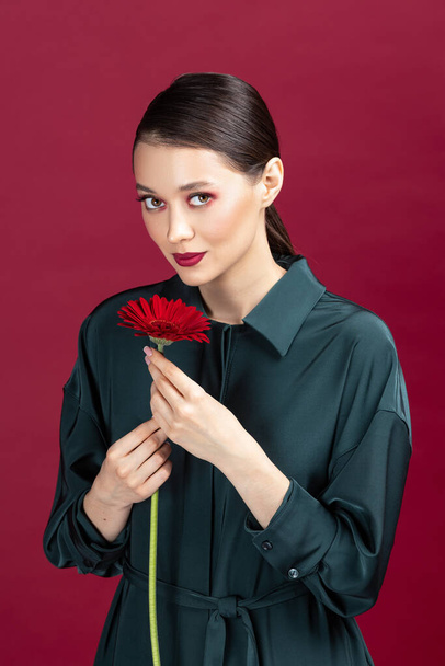 Lovely girl wearing green dress standing isolated over red background, posing with a herbera flower - Zdjęcie, obraz