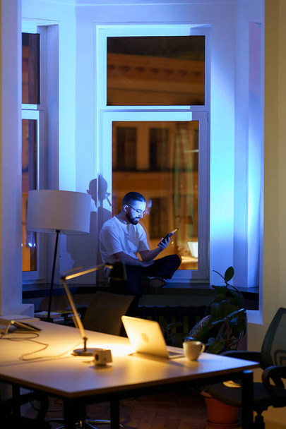 Виснажений бізнесмен відпочиває від роботи понаднормово грати в гру на смартфоні, прокрастинації в темному офісі
 - Фото, зображення
