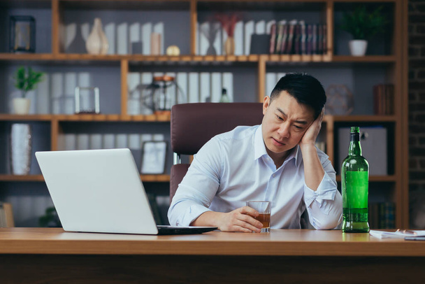 asiático hombre de negocios falla, hombre en desesperación bebe licor duro, sentado tarde en la mesa en la oficina, deprimido y desesperanzado - Foto, Imagen