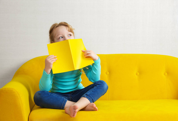 une fille blonde est assise sur le canapé dans la chambre et lit un livre en couverture jaune - Photo, image