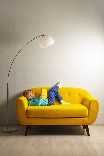 Μια ξανθιά κοπέλα κάθεται στον καναπέ του δωματίου και διαβάζει ένα βιβλίο με κίτρινο εξώφυλλο. - Φωτογραφία, εικόνα