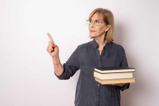 Portret van een oudere blanke vrouw met boeken die met de vinger wijzen naar kant geïsoleerd over een witte achtergrond. - Foto, afbeelding