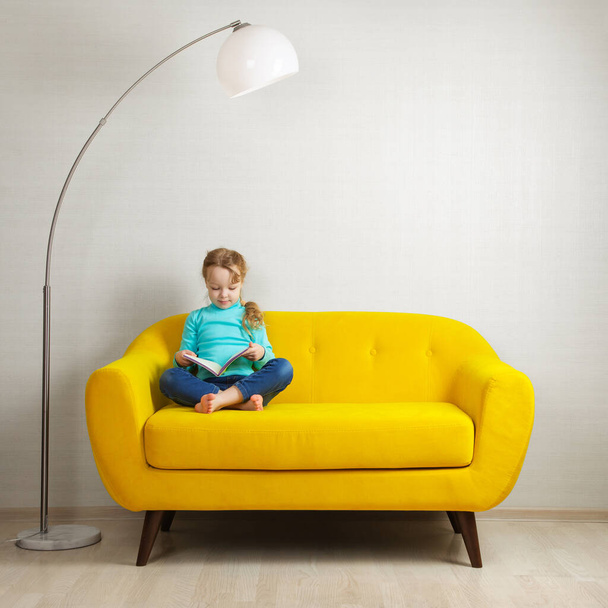 una chica rubia está sentada en el sofá de la habitación y leyendo un libro en una cubierta amarilla - Foto, imagen