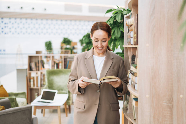 Adult smiling brunette business woman forty years with long hair in stylish beige suit and jeans at public place, green open space office, coworking. Friendly teacher or mentor with book in the library - Foto, imagen