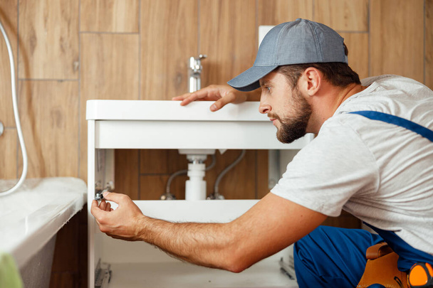 Концентрированный сантехник, мужчина в униформе, чинит раковину и водопровод в ванной комнате - Фото, изображение