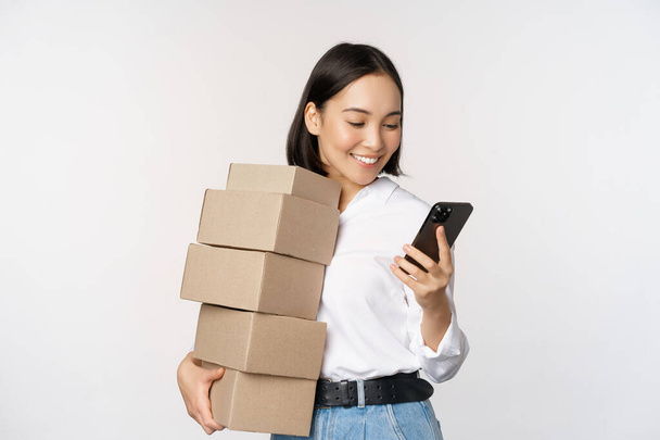 Kuva nuoresta aasialaisesta naisesta, jolla on laatikoita, asiakkaiden tilauksia ja matkapuhelimen katselua, valkoisella taustalla - Valokuva, kuva