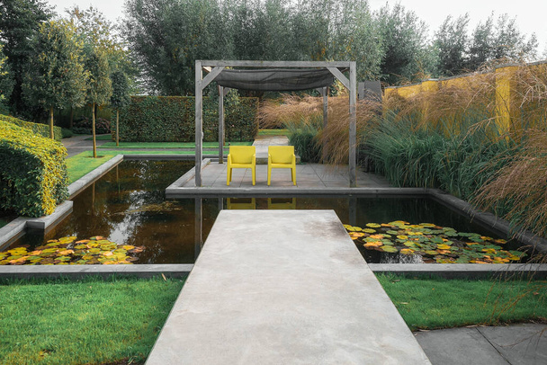 美しい庭の隅に太陽を楽しむために水のプールに囲まれているペルゴラの下に2つの黄色の椅子. - 写真・画像