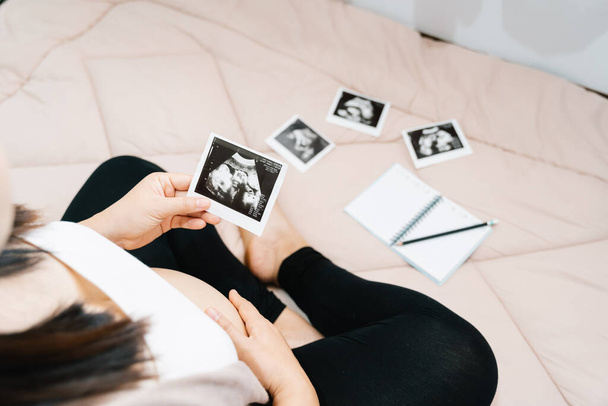 A cute pregnant belly and x ray ultrasound scan of baby. asian young pregnant female relax in the room looking at her child picture in hands sweet. motherhood concept third trimester - Foto, Imagem