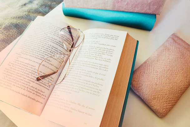 Κοντινό πλάνο. Ρετρό γυαλιά που αναπαύονται στο ανοιχτό βιβλίο, δίπλα στη θήκη ροζ γυαλιών και μερικά βιβλία, στο κομοδίνο στην κρεβατοκάμαρα, το πρωί. - Φωτογραφία, εικόνα