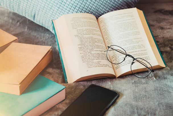 Közepes lövés. Szemüveg pihen a nyitott könyv mellett a párna, a mobil telefon és néhány könyvet az ágyon a hálószobában reggel. - Fotó, kép