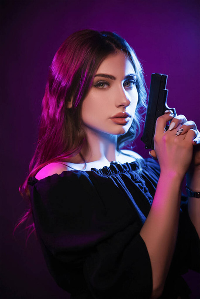 A young woman in the dark in a club holds a gun. Party girl in blue and pink light. Beautiful model portrait in cyber style with guns - Foto, Imagen
