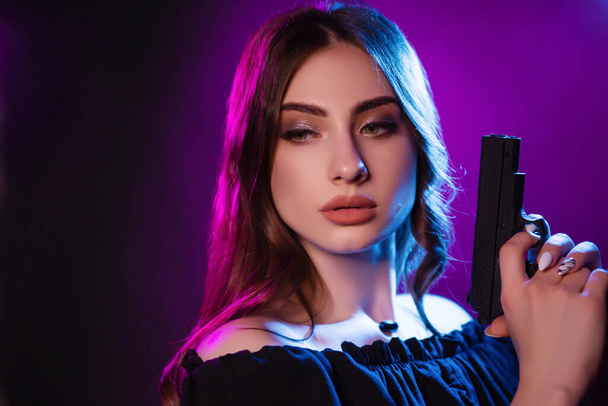 Una joven en la oscuridad en un club sostiene un arma. Chica de fiesta en luz azul y rosa. Hermoso retrato modelo en estilo cibernético con pistolas - Foto, Imagen