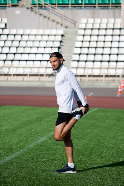 white man in black shorts and a white T-shirt stretches before running on the green lawn of the stadium - Foto, Imagem