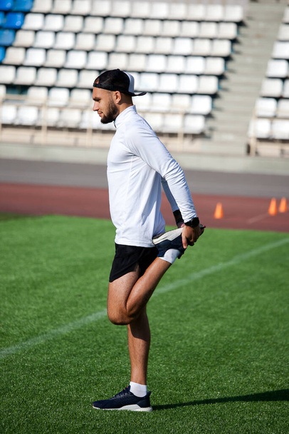 white man in black shorts and a white T-shirt stretches before running on the green lawn of the stadium - Valokuva, kuva