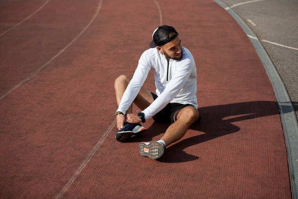 male runner in white black clothes holds his knee at the stadium, sports injury - Fotoğraf, Görsel