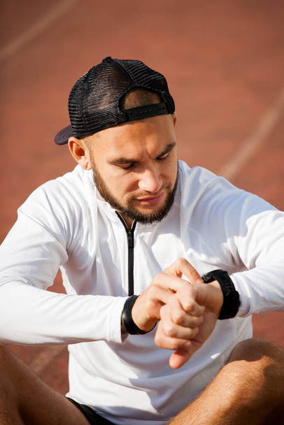 a white man in black shorts and a white shirt looks at a sports watch and adjusts it. Close-up of young man using smartwatch - 写真・画像