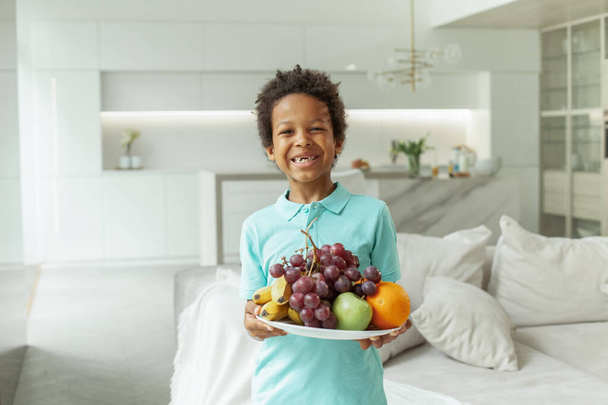 Chłopiec z czarnymi włosami i ładny uśmiech trzymając owoce wewnątrz - Zdjęcie, obraz