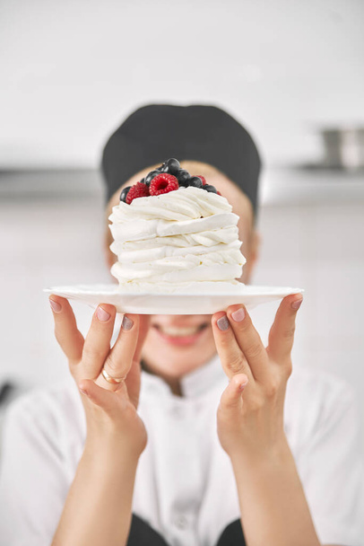 Confectioner in uniform holds a pavlova dessert in his hands, demonstrates in the kitchen. vertical frame, Copy space for text - Zdjęcie, obraz