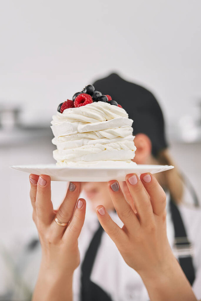 Confectioner in uniform holds a pavlova dessert in his hands, demonstrates in the kitchen. vertical frame, Copy space for text - Photo, image