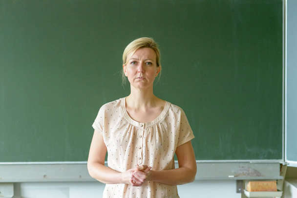 Προνοητικός δάσκαλος του σχολείου συνοφρυωμένος με θλιμμένα μάτια μπροστά από έναν πίνακα σε μια τάξη. - Φωτογραφία, εικόνα