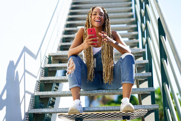 Zwart meisje met gekleurde vlechten, zittend met een smartphone en een skateboard. - Foto, afbeelding
