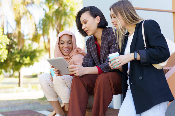 Stylish diverse women friends sharing tablet and drinking takeaway coffee on street - Photo, image