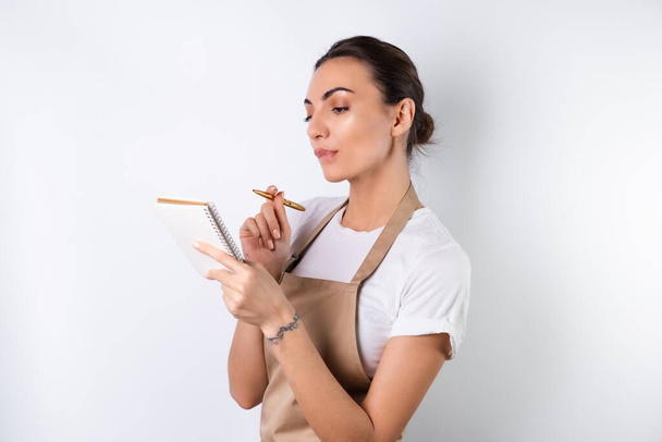 A young housewife in an apron on a white background with a notebook in her hands, chooses recipes for dinner, thinks how to please her husband, comes up with ideas for dishes - Fotoğraf, Görsel