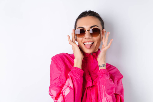 Stylish young woman in a bright pink sports jacket, fashionable sunglasses and wireless headphones in her ears, on a white background shows her tongue - Photo, Image