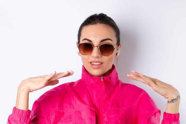Stylish young woman in a bright pink sports jacket, fashionable sunglasses and wireless headphones in her ears, on a white background - Fotó, kép