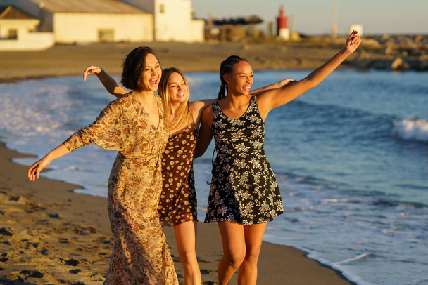 Drie grappige multi-etnische meisjes dragen zomer jurk lachen en plezier hebben samen op het strand. - Foto, afbeelding