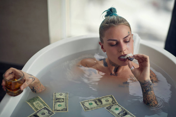 A young beautiful tattooed girl enjoying a cigar and a drink while having a warm bath in the water full of money in a relaxed atmosphere in the bathroom. Relaxation, bath, hedonism, luxury, home - Фото, зображення