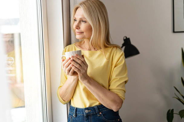 Calm and peaceful middle-aged woman takes a break, rests with a cup of hot tea near the window at home. Contented charming mature lady enjoying coffee - Фото, изображение