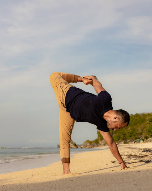 アジア人男性がビーチでヨガを練習しています。運動を伸ばす。柔軟な体にフィット。健康的なライフスタイル。バランスと集中力。セルフケアの概念。ヨガの隠れ家だ。コピースペース。インドネシアのバリのトーマスビーチ - 写真・画像