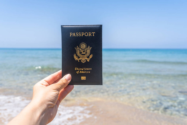 Woman holding american passport in a cover opposite sea wave beach. Concept of summer travel, tourism, holiday. - Zdjęcie, obraz