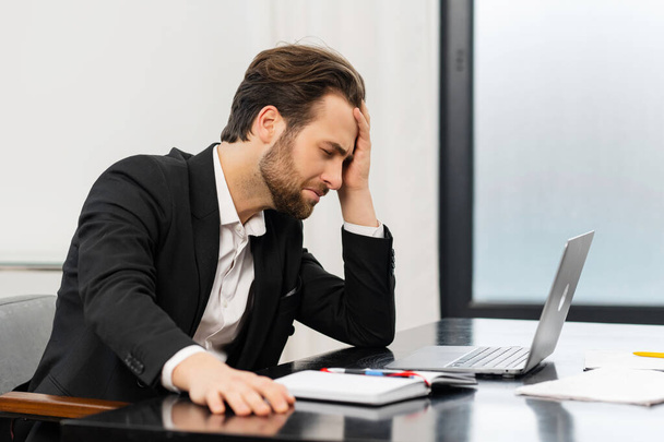Tired businessman frown while checking financial documents, touching forehead with the arm, sitting in the trendy office at the laptop - Foto, Imagen