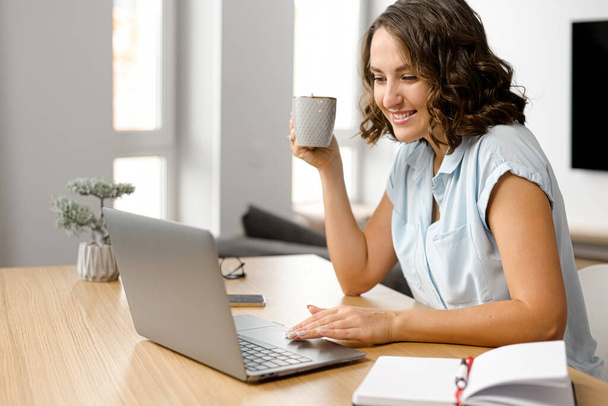 Female freelancer daydreaming with a cup of morning coffee, smiling woman sits at the table with a laptop, looks at the screen - Fotoğraf, Görsel
