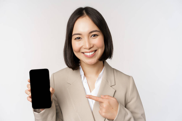 Portrait of corporate asian woman showing smartphone app interface, mobile phone screen, standing over white background - Photo, Image